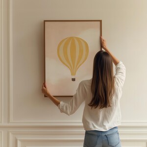 May include: A person hangs a framed print on a white wall. The print features a yellow hot air balloon with white stripes against a pink and white watercolor background.