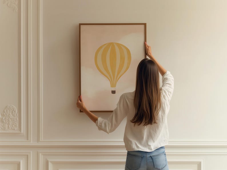 May include: A person hangs a framed print on a white wall. The print features a yellow hot air balloon with white stripes against a pink and white watercolor background.