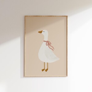 May include: A framed art print featuring a whimsical illustration of a white goose with a pink ribbon around its neck. The goose has a yellow beak and feet, set against a soft beige background. The artwork is enclosed in a light brown frame.