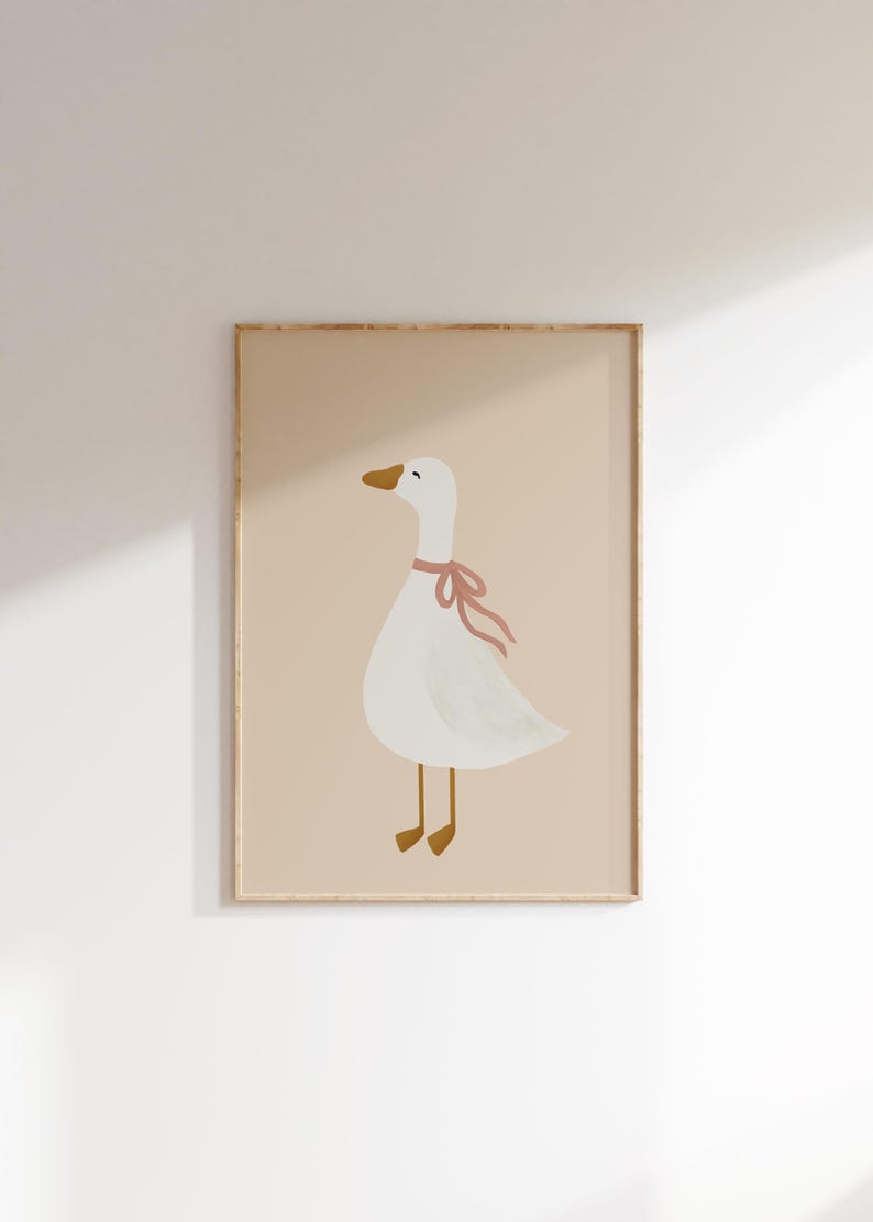 May include: A framed art print featuring a whimsical illustration of a white goose with a pink ribbon around its neck. The goose has a yellow beak and feet, set against a soft beige background. The artwork is enclosed in a light brown frame.