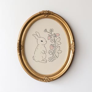 May include: A gold oval frame with a white rabbit and pink strawberries on a cream background.