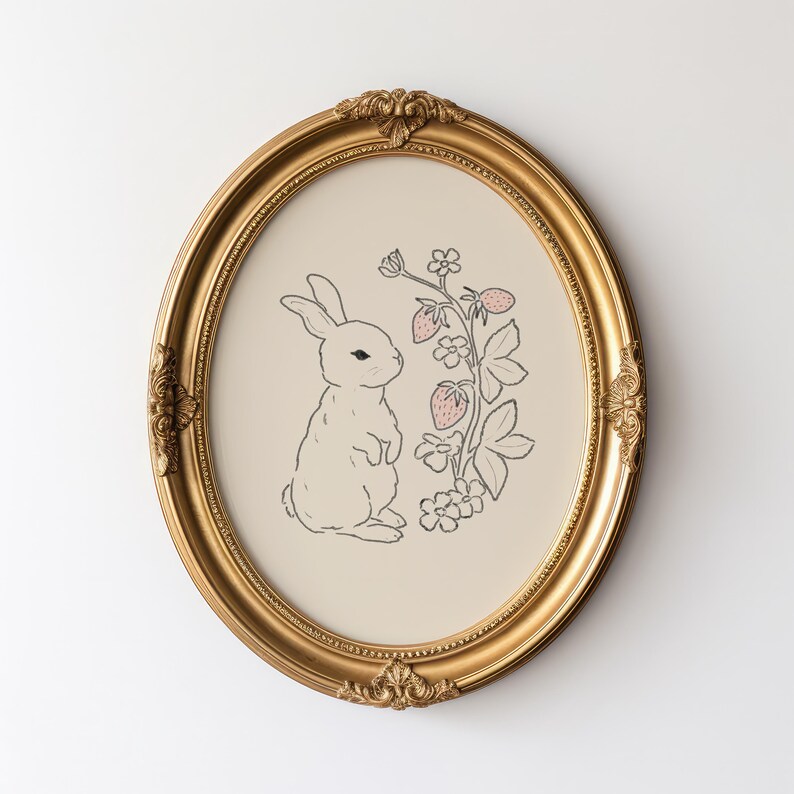 May include: A gold oval frame with a white rabbit and pink strawberries on a cream background.