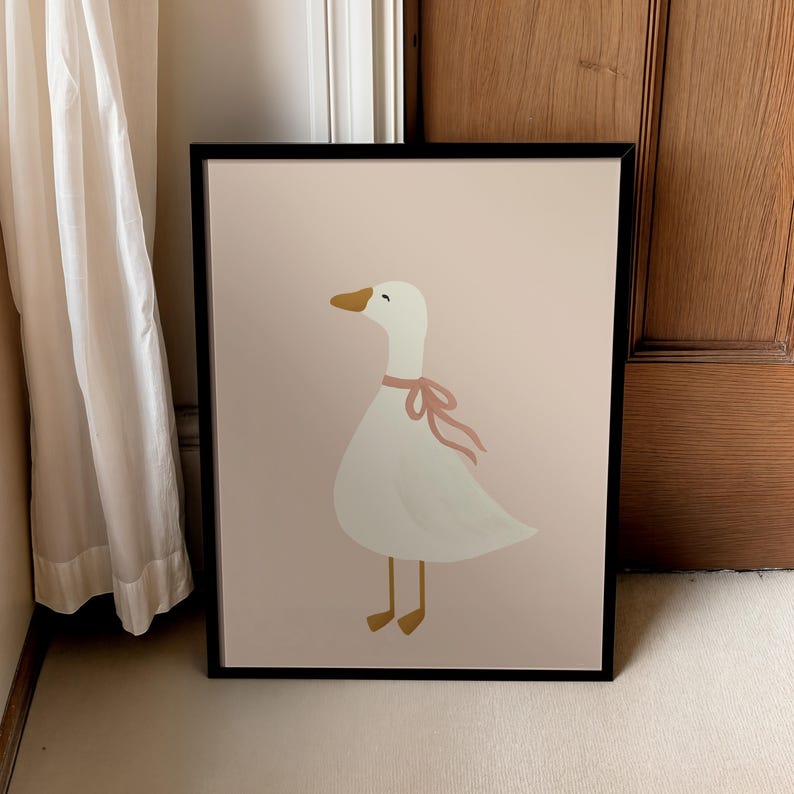 May include: A framed art print featuring a whimsical illustration of a white goose with a pink ribbon around its neck. The goose has a yellow beak and feet, set against a soft pink background. The artwork is enclosed in a black frame.