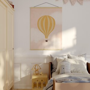 May include: A yellow and white hot air balloon print with a pink background hangs on a wall above a bed with a blue and white checkered blanket. A small wooden lamp sits on a wooden side table next to the bed.