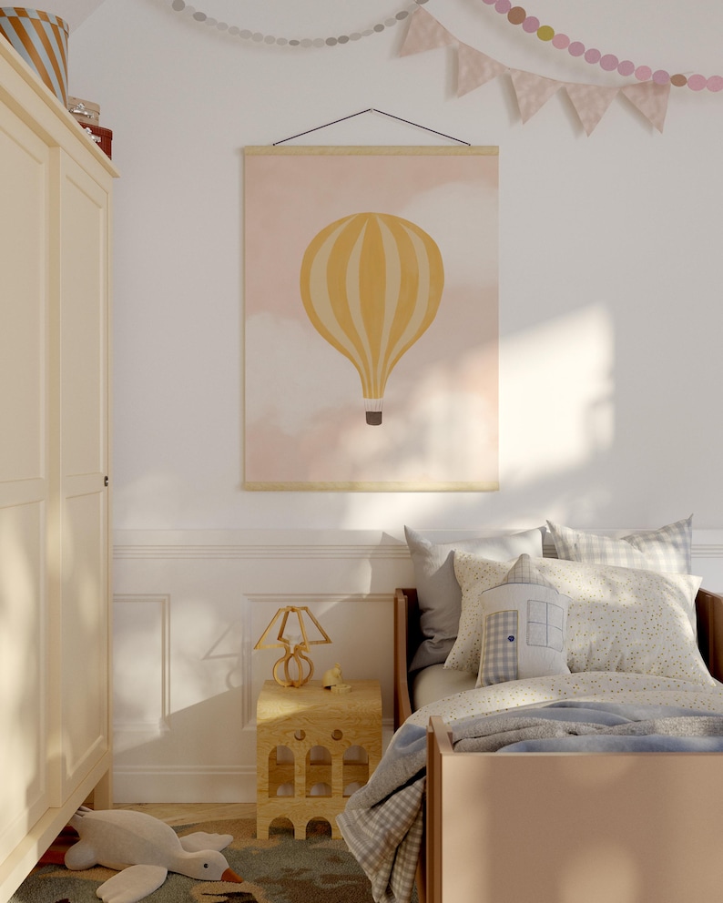 May include: A yellow and white hot air balloon print with a pink background hangs on a wall above a bed with a blue and white checkered blanket. A small wooden lamp sits on a wooden side table next to the bed.