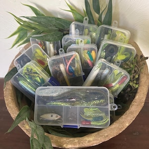 May include: A collection of clear plastic tackle boxes filled with various fishing lures, including soft plastic worms, crankbaits, and hooks. The boxes are arranged in a decorative, textured bowl with green foliage accents, creating a visually appealing display for anglers.