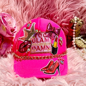 May include: A bright pink trucker hat with a gold chain and patches. Patches include a crown, lips, lipstick, high heels, and the text "Too Glam To Give A Damn". Rhinestones and a sequined bow add sparkle.