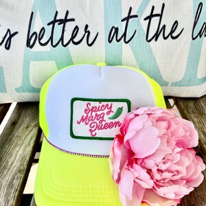 May include: A neon yellow and white trucker hat with the text "Spicy Marg Queen" on a patch. A pink flower rests on the hat. A pillow with the text "is better at the la" is in the background.