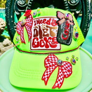 May include: A neon green baseball cap featuring a central patch with the text "I need a Diet Coke" in red and brown. The cap is decorated with two red and white checkered bows, a black sequined soda can patch, and various iridescent rhinestones.