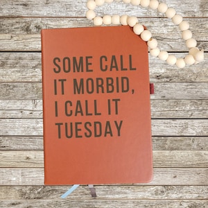 Op de afbeelding: Een roestkleurig notitieboek met de zwarte tekst "SOME CALL IT MORBID, I CALL IT TUESDAY" op de voorkant. Een snoer van lichtgekleurde houten kralen ligt bovenop het notitieboek en twee kleine lint-bladwijzers steken aan de onderkant uit.