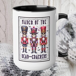 May include: White ceramic mug with a black interior and handle. The mug features a rectangular design with the text "MARCH OF THE DEAD-CRACKERS" and illustrations of three skeleton nutcrackers in colorful uniforms. The mug is held by a person wearing white and gray mittens.