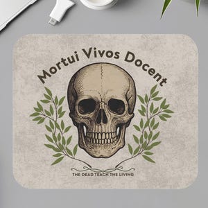 Op de afbeelding: Een rechthoekige muismat met een schedelillustratie, de Latijnse zin "Mortui Vivos Docent" en de Engelse vertaling "The Dead Teach the Living". Het ontwerp toont een gedetailleerde schedel met olijftakken.