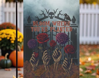 May include: A rectangular garden flag featuring a dark, atmospheric scene of four skeletal hands emerging from the soil, each holding a vibrant red or purple flower. Above the hands, the text BLOOM WHERE YOU'RE PLANTED is displayed in red.