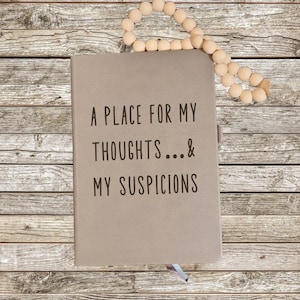 May include: A tan journal with the text "A PLACE FOR MY THOUGHTS...& MY SUSPICIONS" in black. A beaded garland rests above the journal on a wooden surface. A ribbon bookmark is visible.