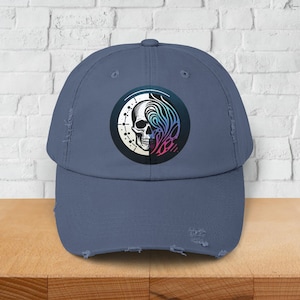Op de afbeelding: Een blauwe baseballcap met een distressed look, met een cirkelvormig ontwerp met een schedel en abstracte kleurrijke patronen. De cap heeft een voorgewassen look met gerafelde randen.