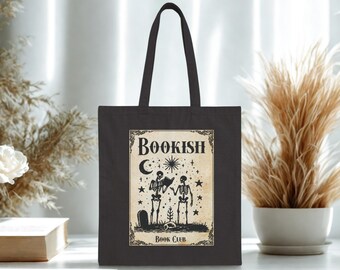 Puede incluir: Bolso tote negro con un diseño rectangular que presenta dos figuras de esqueleto leyendo libros bajo una luna creciente y estrellas. El texto "Bookish Book Club" se muestra en el diseño.