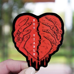 May include: A red heart-shaped sticker with a stitched split down the middle. The left side features a human hand, while the right side shows a skeletal hand. The words "NOT VIABLE" are vertically printed on the left side of the heart. Black dripping blood detail.