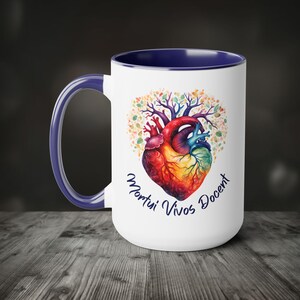 May include: White ceramic mug with a blue handle and interior. The mug features a colourful anatomical heart design with a tree-like pattern and the text "Mortui Vivos Docent" in blue script. The mug is on a wooden surface.