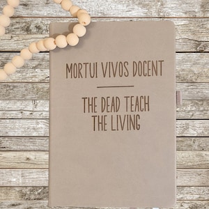Op de afbeelding: Een beige notitieboek met de gegraveerde tekst "MORTUI VIVOS DOCENT" en "THE DEAD TEACH THE LIVING". Een kralenkrans bevindt zich in de linkerbovenhoek. Het notitieboek heeft een lint bladwijzer.