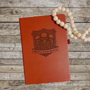 May include: A terracotta-coloured journal featuring a debossed skull design and the Latin phrase "Mortui Vivos Docent." A strand of cream-coloured wooden beads rests on the wooden surface next to the journal.