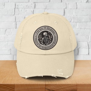 Op de afbeelding: Crèmekleurige distressed baseballcap met een cirkelvormig embleem met een skelet en de tekst "I've Got 99 Problems & They're All Unidentified Remains." De cap heeft een gebogen klep en een versleten uiterlijk.