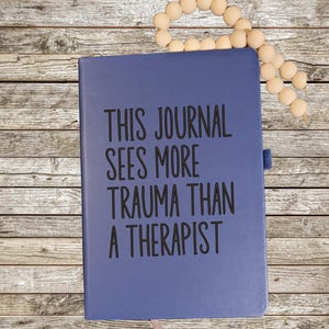 May include: A blue journal with the text "THIS JOURNAL SEES MORE TRAUMA THAN A THERAPIST" in black. A light pink ribbon bookmark and a strand of wooden beads are also visible.