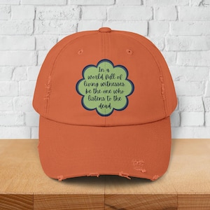 Op de afbeelding: Een versleten oranje baseballpet met een groene, bloemvormige patch waarop staat: "In een wereld vol levende getuigen. Wees degene die naar de doden luistert."