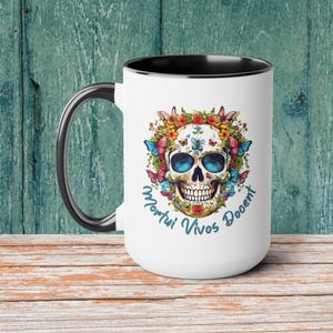 May include: White ceramic mug with a black handle and interior. The mug features a colorful skull design with butterflies, flowers, and the text "Mortui Vivos Docent" in blue script. The mug is set against a teal wooden background.