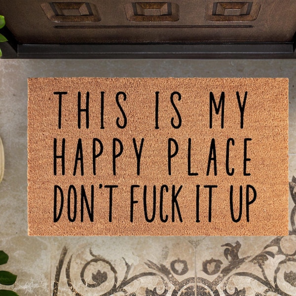 May include: A rectangular doormat made of natural coir fiber. The mat features the text "THIS IS MY HAPPY PLACE DON'T FUCK IT UP" in black, bold letters. The mat is placed in front of a door.