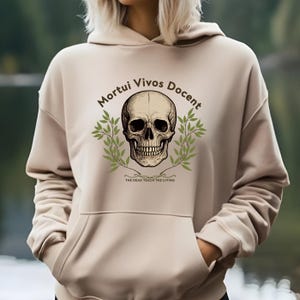 Könnte beinhalten: Beigefarbener Kapuzenpullover mit Fronttasche und Kordelzugkapuze. Auf der Vorderseite befindet sich eine detaillierte Totenkopf-Grafik, umgeben von einem grünen Kranz, mit dem Text "Mortui Vivos Docent" und "The Dead Teach The Living".