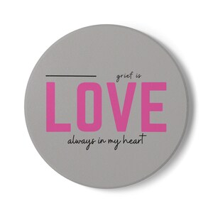 May include: A circular gray coaster featuring the text "grief is" in small black letters above the word "LOVE" in large, bold pink capital letters, with a black horizontal line to the left and "always in my heart" in black script below.