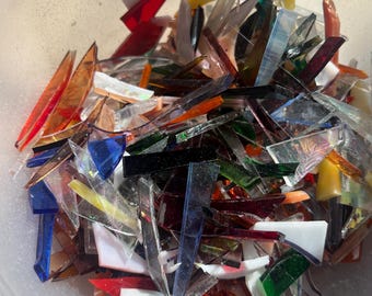 May include: A clear plastic container filled with a variety of colorful, irregularly shaped glass pieces. The glass shards are in shades of red, orange, yellow, green, blue, and white. The pieces vary in size and shape, with some appearing sharp and pointed.