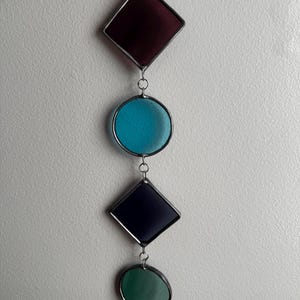 May include: A vertical hanging ornament featuring four geometric stained glass pieces connected by metal jump rings. The shapes include a deep red diamond, a bright turquoise circle, a dark navy blue diamond, and a forest green circle.