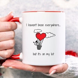 May include: White ceramic mug with a red rim and handle. The mug features a black and white illustration of a hot air balloon shaped like a heart with two stick figures inside. The text on the mug reads "I haven't been everywhere... but it's on my list."