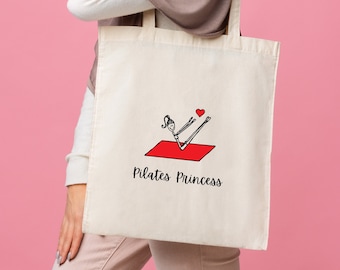 Può includere: Una borsa tote bianca con un'illustrazione rossa e nera di una persona che fa pilates su un tappetino rosso. Il testo "Pilates Princess" è stampato sotto l'illustrazione.
