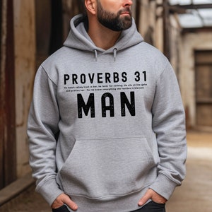 Pode incluir: Uma camisola de capuz cinzenta com o texto "PROVERBS 31" e "MAN" em preto. O texto diz "O seu coração confia nela com segurança, ele não precisa de nada. Ele senta-se à porta e a elogia. Porque ele sabe que tudo o que ela toca é abençoado."