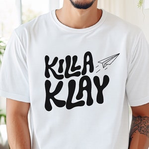 May include: White t-shirt with black text that reads "KILLA KLAY" and a paper aeroplane graphic.