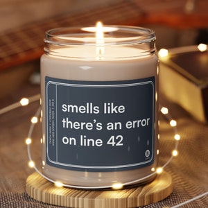 以下が含まれることがあります： 白いラベルに「smells like there's an error on line 42」と書かれた茶色の大豆ワックスキャンドル。キャンドルは木製のコースターの上に置かれています。