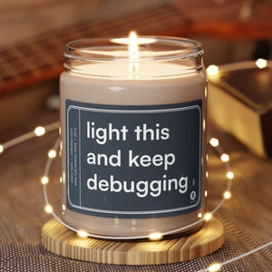May include: A brown soy wax candle with a white label that reads "light this and keep debugging". The candle is lit and sitting on a wooden coaster.