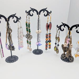May include: A display of eight pairs of dangle earrings with various beads, charms, and colors. The earrings are displayed on a black metal stand.