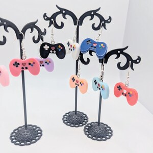 May include: A set of six colorful video game controller earrings. The earrings are displayed on a black earring stand. The controllers are pink, blue, white, and black.