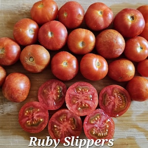 May include: A collection of red Ruby Slippers tomatoes, some halved, arranged on a wooden cutting board. An orange-handled knife rests near the tomatoes. The text "Ruby Slippers" is at the bottom.