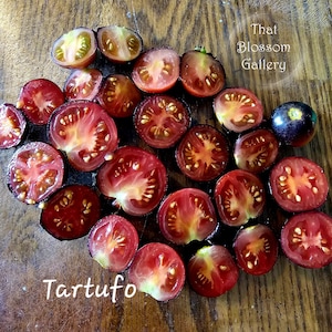 May include: A collection of halved and whole Tartufo tomatoes, showcasing their deep red and purple hues. The cut tomatoes reveal a vibrant interior with visible seeds, arranged on a wooden surface. Text reads "That Blossom Gallery" and "Tartufo".