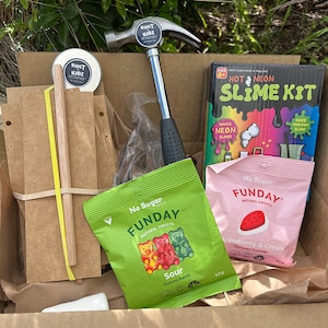 May include: A brown cardboard box filled with various items, including a hammer, a wooden dowel, a packet of sour gummy bears, a packet of strawberry and cream gummies, and a slime kit. The box also contains two small round metal containers with the text "Built by Kidz Activities" printed on them.