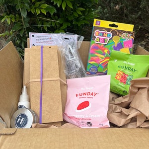 May include: A cardboard box filled with various items, including a craft kit, a bag of sand art, and two bags of gummy sweets. The craft kit includes a wooden board, a bottle of glue, and a small jar of glitter. The sand art kit includes a plastic tray, a funnel, and a variety of coloured sands. The gummy sweets are in bags labelled "No Sugar Funday" and feature strawberry and cream and sour gummy bears.