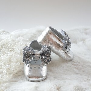 May include: A pair of silver baby shoes with a sparkly bow on each shoe. The shoes are on a white fur surface.