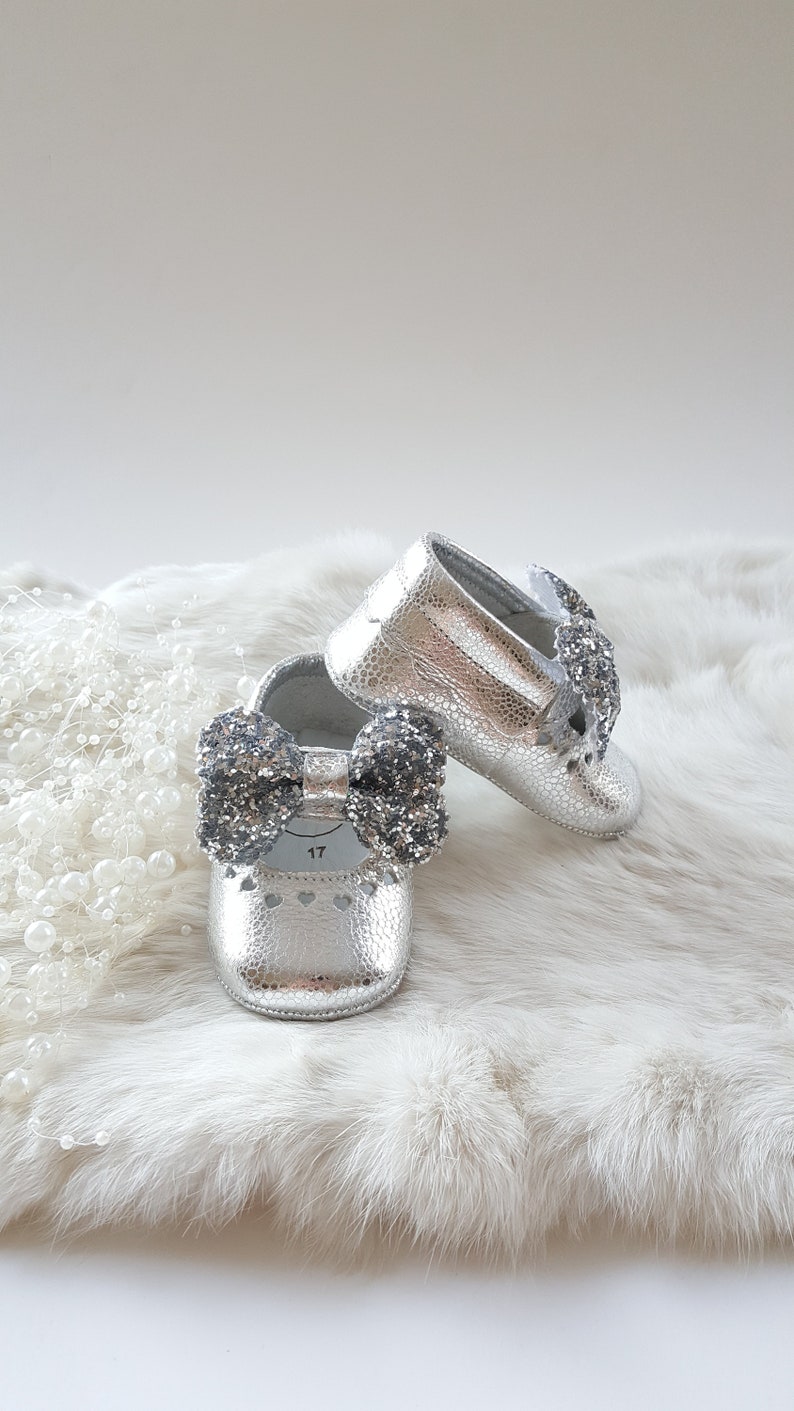 May include: A pair of silver baby shoes with a sparkly bow on each shoe. The shoes are on a white fur surface.