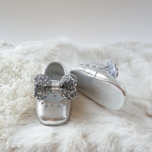 May include: A pair of silver baby shoes with a sparkly bow on the front. The shoes are on a white fur background.