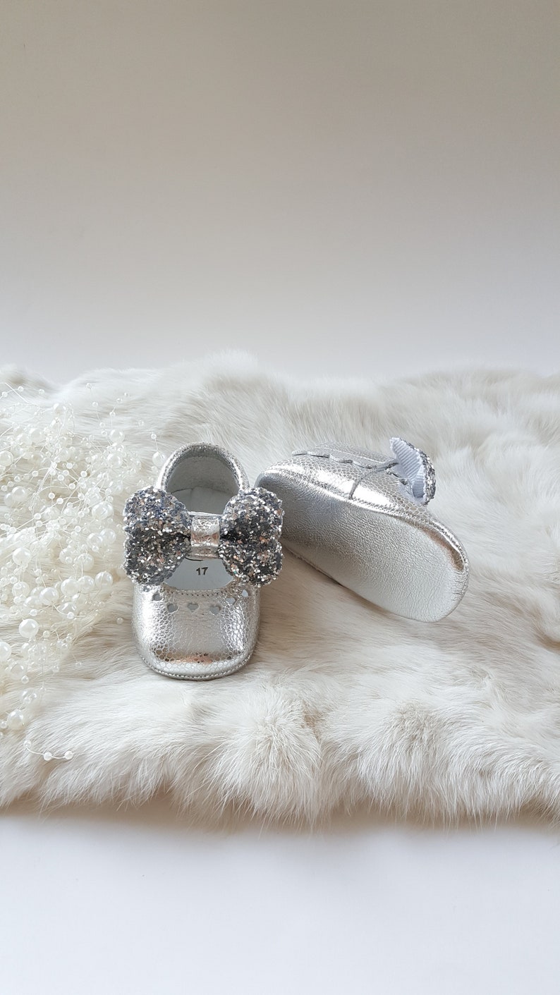 May include: A pair of silver baby shoes with a sparkly bow on the front. The shoes are on a white fur background.