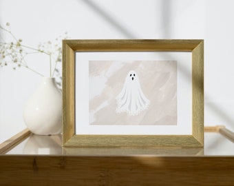 May include: A framed print of a white ghost illustration with a lace trim. The artwork is set against a neutral background and framed in a gold-coloured frame. The print is displayed on a glass-topped table.